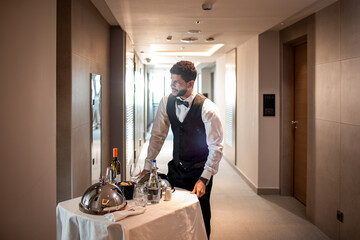 Young waiter delivering tray with food in a room of hotel doing room service