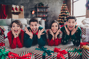 Fototapeta premium Full body portrait of group impressed positive fellows lying floor raise hands open mouth christmas giftbox decorated house indoors