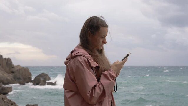 Young Woman in a Storm by the Sea Stands on the Rocks, Looking at Something on Her Phone. Overcast weather, she wears a Pink Raincoat.