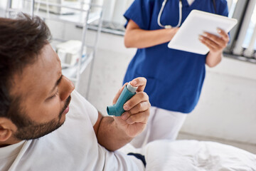 cropped view of indian man using asthma inhaler with blurred nurse on backdrop, healthcare