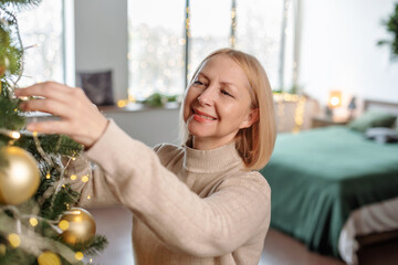Mature woman decorate Christmas tree of ornaments and garland lights.
