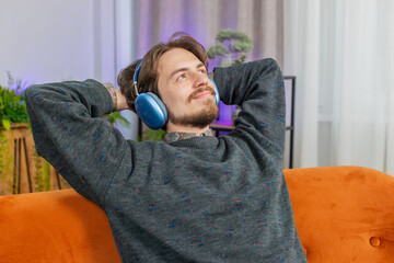 Happy smiling Caucasian young man in wireless headphones relaxing chill lying on home couch choosing listening favorite energetic disco rock n roll music entertaining. People weekend activities