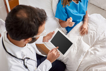 african american patient sitting on bed near her indian doctor taking notes on tablet, cropped