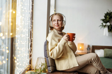 Mature woman sit near at window and looks from Christmas room, decorations garland lights. Holiday...