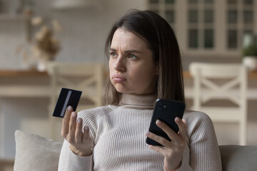 Frustrated young woman sit on sofa hold credit card use phone try to pay through e-bank app, experiences problem with e-payment due insufficient funds, no money, scam, unsuccessful transaction concept - obrazy, fototapety, plakaty