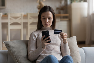 Serious woman sit on sofa holding cellphone and credit card buy goods make order on internet use retail e-services. Secure payment through e-bank app, instant money transfer, e-commerce client concept