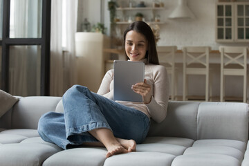 Young pretty smiling woman spend leisure at home rest sit on cozy sofa using digital tablet....
