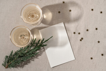 Minimal aesthetic holiday template, blank paper card mockup, wineglasses with sparkling wine, gold confetti and juniper branch on beige linen background, top view, flat lay