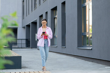 Pleasant young woman in casual pink shirt standing on city street with modern smartphone in hands. Positive brunette typing messages on cell phone outdoors.