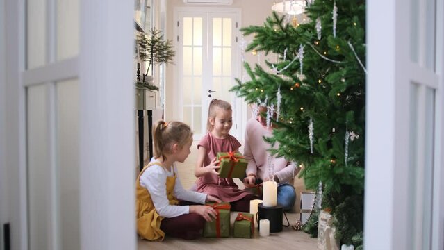 A happy mom with two little daughters is packing Christmas presents sitting under the Christmas tree on the floor in the living room