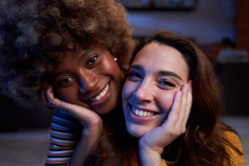 Cozy portrait of a young multiracial LGBTQ couple lying on the floor at home, looking at camera smiling. Close up of two cheerful woman indoors together at night. - obrazy, fototapety, plakaty