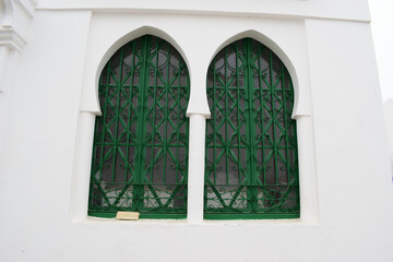 Two arched windows with green decorative metal grilles on a white wall, Islamic architecture, symmetrical design, clean lines, historic building, exterior detail, cultural heritage.