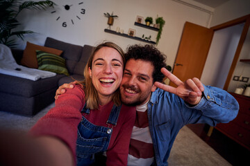 Cheerful couple at home using smartphone taking selfie to post on social media, having fun together in new apartment. POV people lifestyle. - obrazy, fototapety, plakaty
