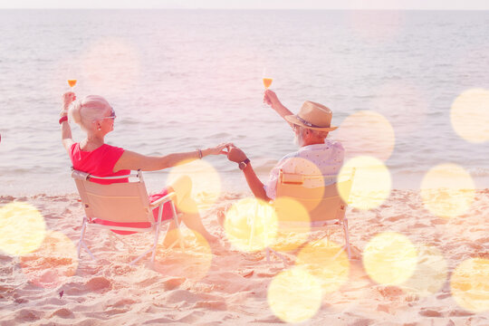 Senior couple celebration : Back of senior man holding juice and sheers to his wife during sit on beach chair and relax on holiday or vacation time with celebration bokeh light.