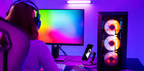 Asian professional gamer playing online video game on desktop computer PC have colorful neon LED lights, young woman in gaming headphones using computer for playing game at home, back view