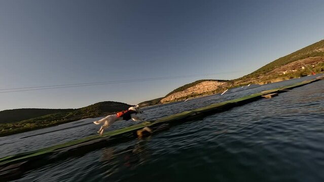 A white dog dressed in a life jacket quickly runs across the bridge after the owner who rides a wakeboard in a wakepark. High quality 4k footage fpv drone shoot sunset a lake surrounded by mountains