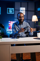 African american man filming video for online channel in dimly lit home studio, presenting gaming headphones to subscribers. Content creator showing viewers audio listening device