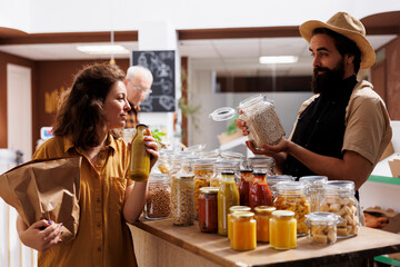 Woman shopping in zero waste grocery store, looking at bulk products in reusable packaging while being assisted by friendly vendor. Customer in local neighborhood picking locally sourced food