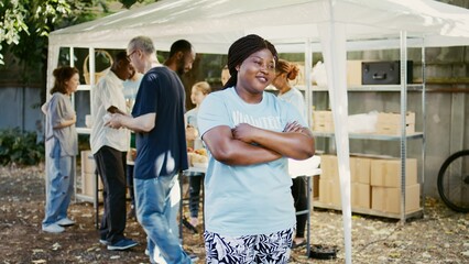 Enthusiastic black female volunteer, ready to provide humanitarian aid at a charity food distribution event. Multiethnic charity workers handing out free food to homeless people. Side-view, portrait. - obrazy, fototapety, plakaty