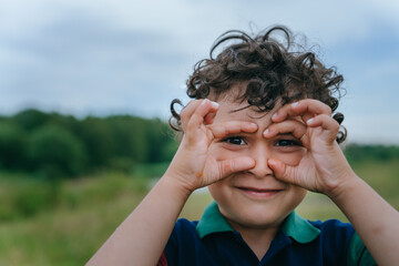 Curly hispanic little boy in polo stands on meadow, against forest makes binoculars by hands looks...