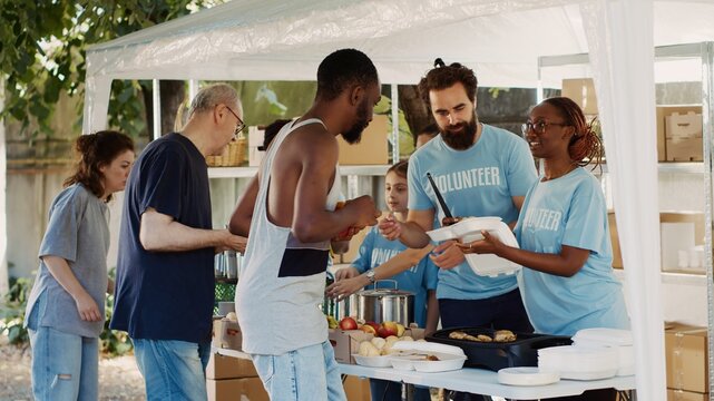 Humanitarian aid team graciously providing free warm meals to the poor needy and less fortunate individuals. Group of volunteers handing out food donations to the homeless people. Tripod shot.