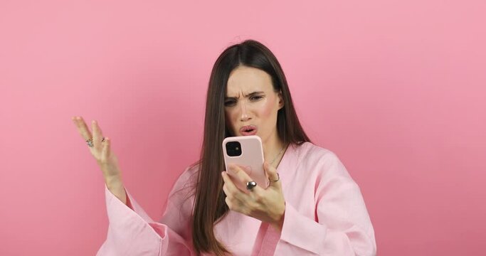Mad and shock young brunette beautiful woman using mobile phone isolated on pink background. Yelling unhappy woman hold mobile phone. Disappointed sad upset lady horrified impressed news. WTF. Oh no