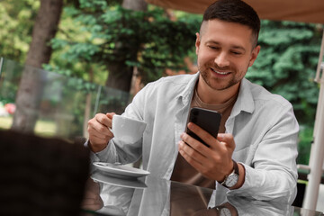 Handsome man with cup of drink sending message via smartphone at table in outdoor cafe