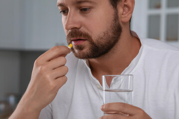 Depressed man with glass of water taking antidepressant pill indoors, closeup