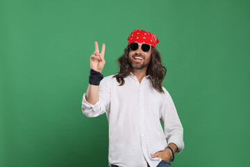 Stylish hippie man in sunglasses showing V-sign on green background