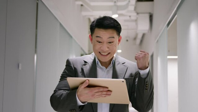 Happy excited middle-aged Asian businessman chinese korean japanese man with digital tablet win business success bet money lottery amazed 40s male winning financial prize achievement at office hall
