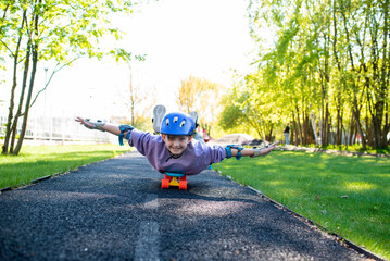 a boy rides on a skateboard lying on his stomach, a child in a purple helmet and protection on his arms and legs lies on a city cruiser in a skate park - obrazy, fototapety, plakaty