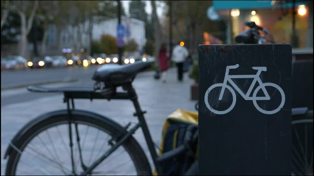 Retro style bicycle parked at the city bike parking lot during the evening. Bike parking metal rack stand with the bicycle icon, on the blurred background with walking people and cars.