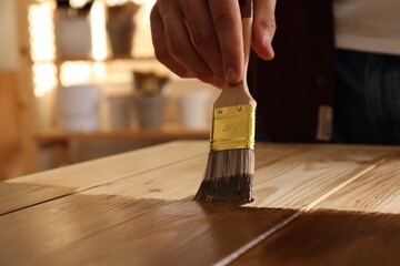 Man with brush applying wood stain onto wooden surface indoors, closeup - obrazy, fototapety, plakaty