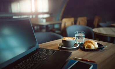Close-up shot, coffree cup with computer laptop and bread for breakfast on wooden table in cafe. vintage light and blurred background