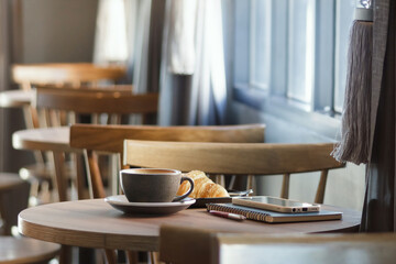 Coffree cup with smart phone placed on a notebook and bread on the plate on wooden table by the window with sunlight in the morning.