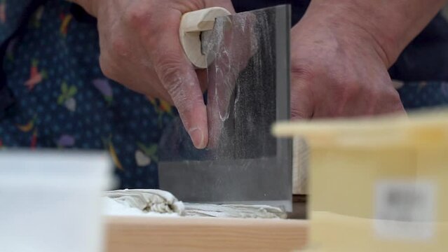 A close-up of a skilled hand meticulously slicing fresh soba noodles with a traditional Japanese knife highlights this culinary art's intricate details and craftsmanship.  Zoom Out.