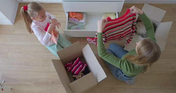 Little girl with mother putting clothes to cardbox in bedroom for donation