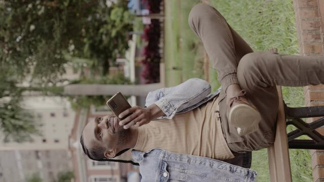 Jovem negro, sentado no banco da praca, falando em seu smartphone. Cinematico 4k. Video vertical.