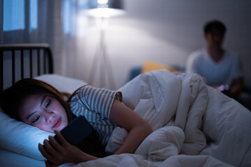 Asian girlfriend reading cartoon before watching Korean drama series while lie down and turn back in bed because sulk and angry boyfriend check online work on a sofa when bedtime night not go to sleep - obrazy, fototapety, plakaty