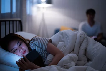Fotobehang Asian girlfriend reading cartoon before watching Korean drama series while lie down and turn back in bed because sulk and angry boyfriend check online work on a sofa when bedtime night not go to sleep © MergeIdea
