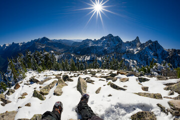 Adventurous athletic male hiker, sitting in a snowy boulder field with his legs outstretched before jagged snowy mountains on a beautiful sunny day.