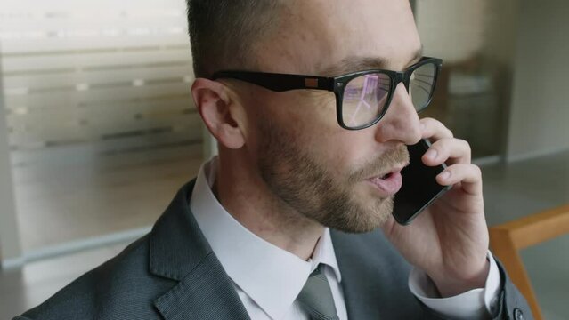 Close up slowmo shot of Caucasian male manager with braces and glasses speaking on phone with business partners in modern office foyer