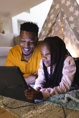 Happy african american father and daughter lying by teepee with tablet at home, copy space