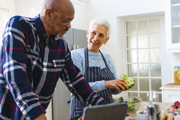 Diverse senior couple preparing healthy meal with vegetables using tablet in kitchen