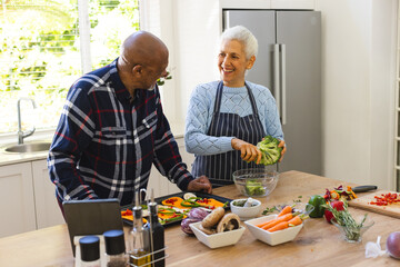 Diverse senior couple preparing healthy meal with vegetables using tablet in kitchen - obrazy, fototapety, plakaty