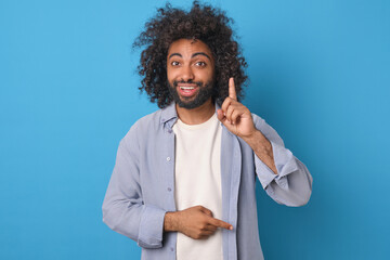 Young cheerful curly Arabian man points finger up indicating presence of new idea for launching...