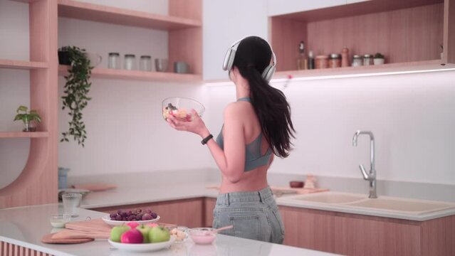 Asian sportswoman enjoying a salad, listening to music, and dancing in her kitchen for fun. She maintains good health with nutritious meals, promoting a positive body image.