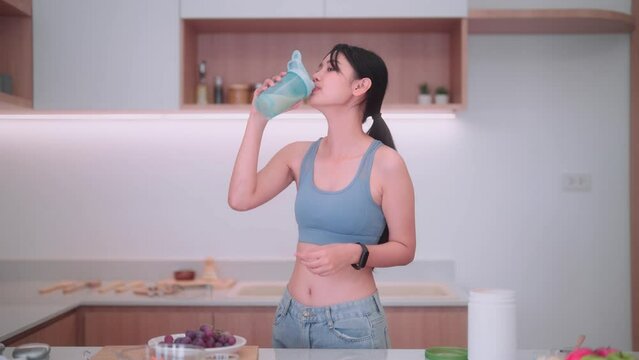 Asian sportswoman in sportswear having a protein shake to manage weight in her home kitchen. She focuses on maintaining good health, consuming nutritious food, and keeping a positive body image.