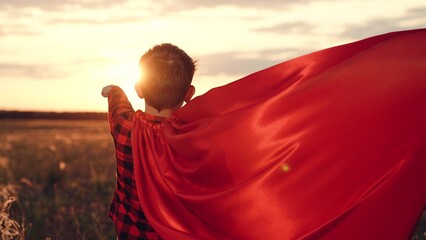 Playful boy stands in superhero character pose with red cape in field at sunset