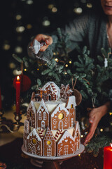 Beautiful woman in green dress making handmade homemade delicious gingerbread house for family Christmas celebration. Elegant table setting. Cozy home atmosphere, festive mood, candlelight, fir tree
